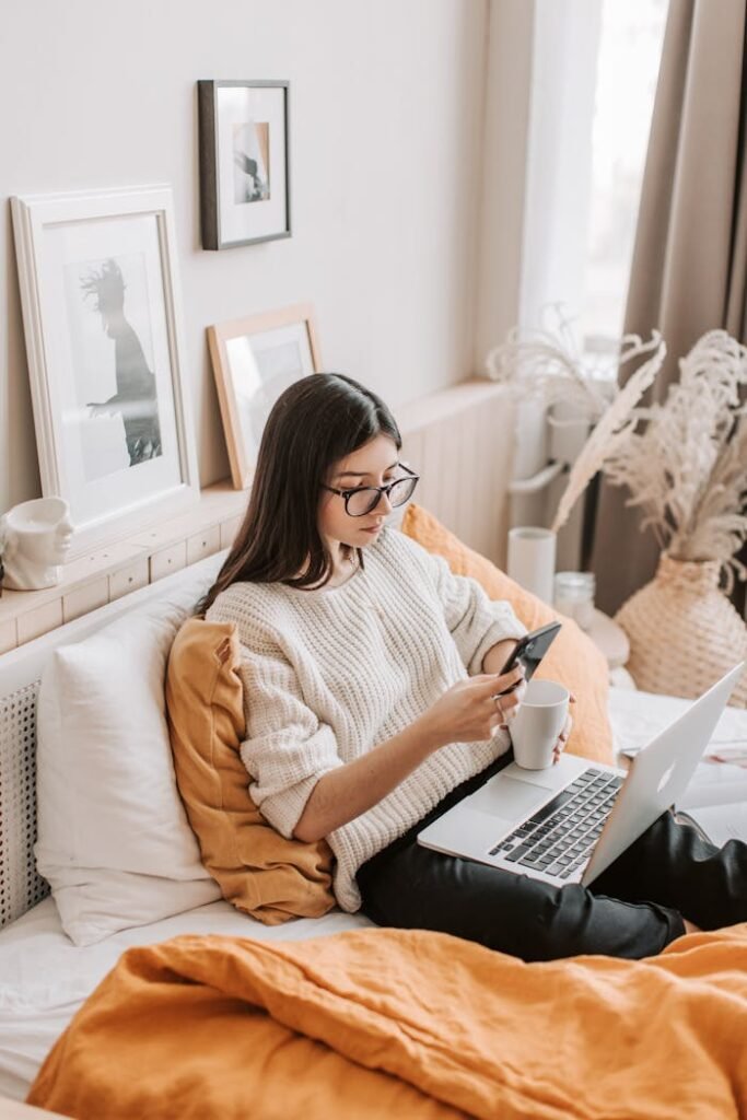 From above of thoughtful young female in warm casual clothes and eyeglasses browsing smartphone while resting on comfortable bed with laptop and cup of hot beverage in daylight